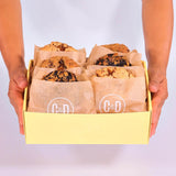 Hands holding 6 x New York Cookies, The Caramel Box by Crumbs & Doilies in a yellow box with branded wrapping.